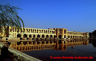 Isfahan - Khadju-Brücke. "Die rund 150 m lange und 14 m breite Pol-e Khadju ..., eine architektonisch sehr reizvolle Brücke mit zweigeschossigen Bogenreihen, wurde um 1650 in der Zeit von Shah Abbas II. erbaut. Ihre 23 Bögen des unteren Geschosses ruhen auf zum Fluß hin abgetreppten, steinernen Fundamenten, die mit Schleusentoren ausgestattet waren. Bei Bedarf konnte so das Wasser des Zayandehrud aufgestaut werden, um die zahlreichen Gartenanlagen des safawidischen Isfahan zu bewässern. Den relativ schmalen Gehweg der Khadju-Brücke im zweiten Geschoß säumen zu beiden Seiten nach außen offene, untereinander verbundene Arkadengänge, deren Bögen mit geometrischem Fliesendekor verziert sind. In der Mitte und ebenso an beiden Enden der Brücke wurden Pavillonbauten in das Bauwerk eingefügt, deren dreiseitige Fassaden ins Wasser hineinragen. Jede der drei Pavillonseiten öffnet sich in einem mit zarten Wandmalereien geschmückten Iwan auf den Fluß. Im mittleren Pavillon, der auch als Shahneshin bezeichnet wird, saßen in safawidischer Zeit der Shah und die Höflinge, um sportlichen Ereignissen oder Wasserspielen auf dem Fluß zuzusehen." Quelle: M. Rashad: Iran. 294. Ostfildern 2006.